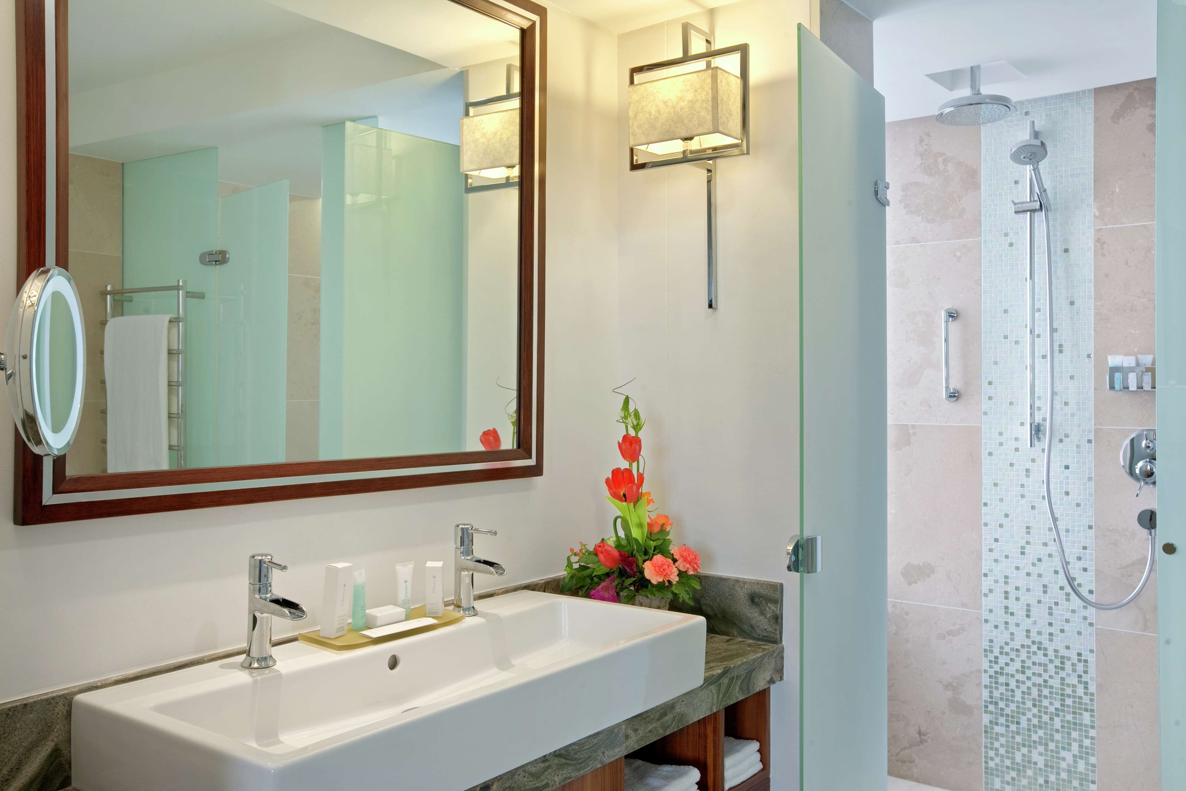 Un baño con un espejo grande, un lavamanos con encimera de mármol, una ducha con puerta de cristal y una lámpara de pared.