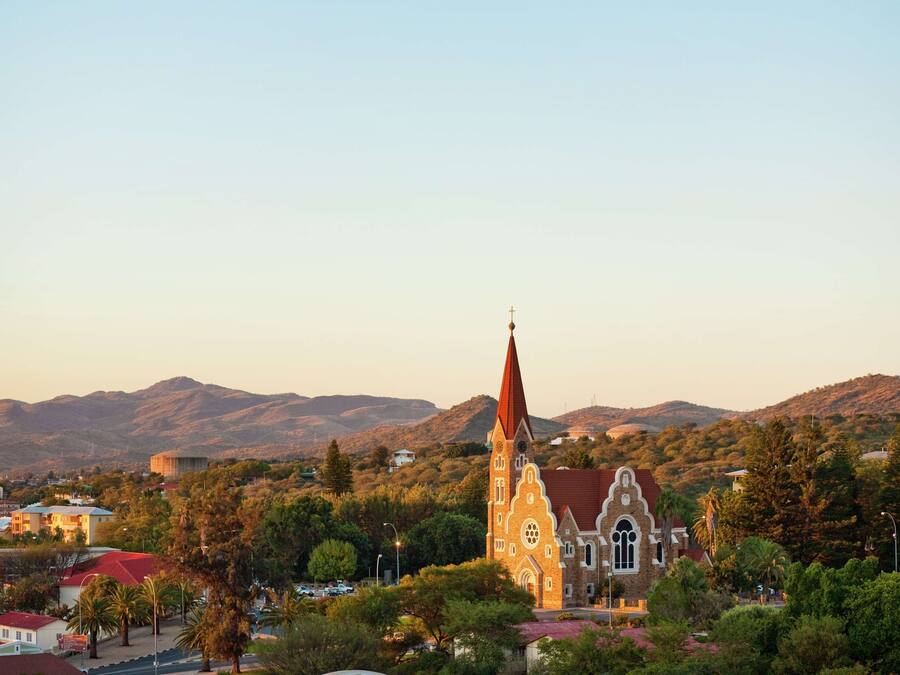 A church with a tall spire, surrounded by trees and buildings, with mountains in the background.