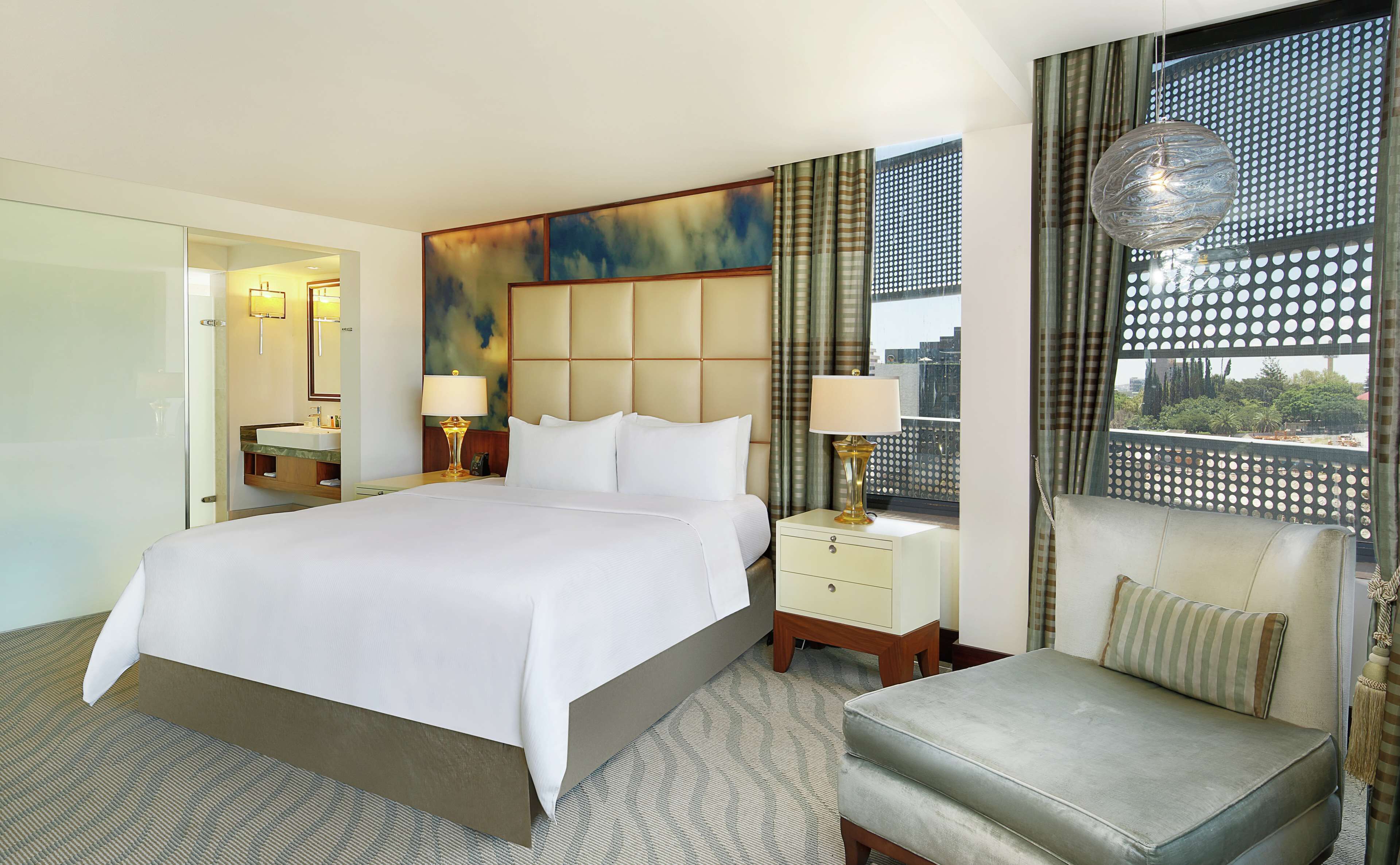 Quarto de hotel com uma cama grande, uma mesinha de cabeceira com abajur, uma cadeira e vista da cidade através de uma janela com cortina estampada.