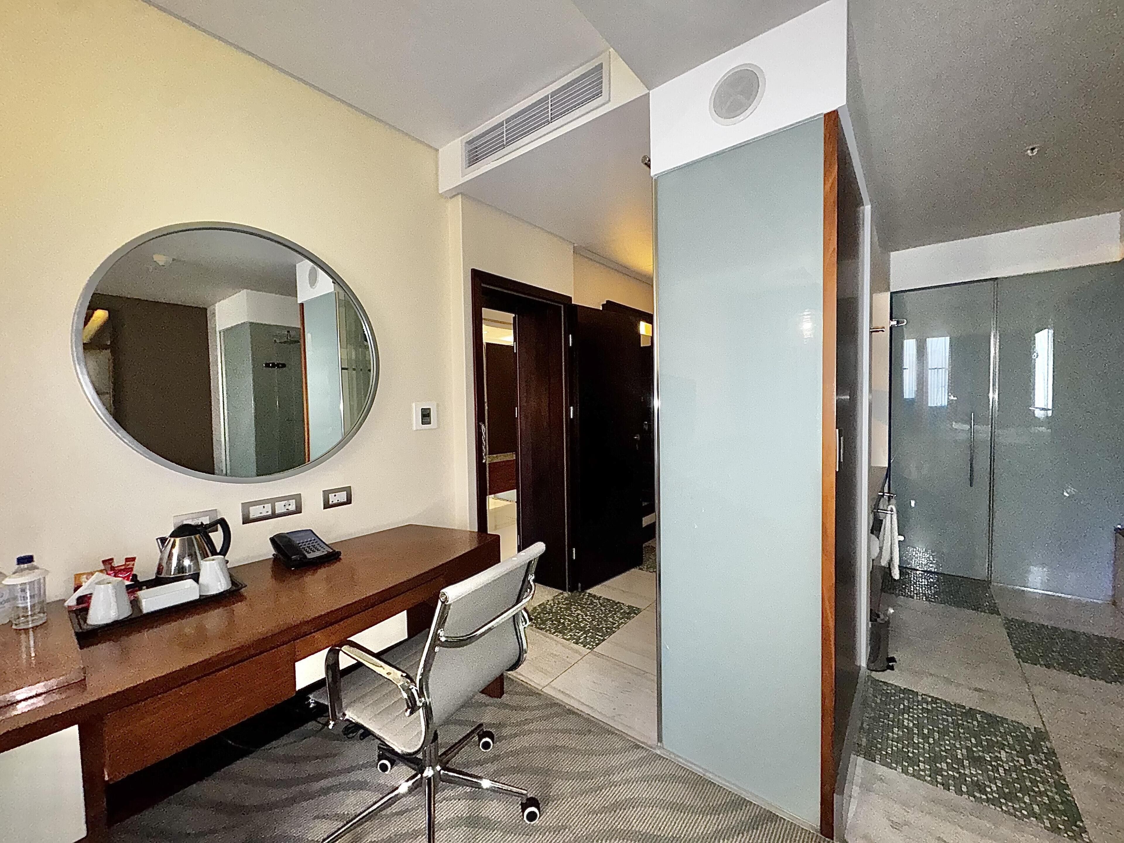 Habitación de hotel con escritorio de madera, un espejo redondo, una silla, zona de ducha y una puerta que da a otra habitación.