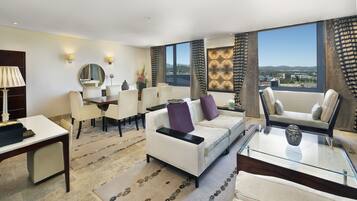 A modern hotel room with a dining area, a sofa set, a glass coffee table, and large windows offering a city view.
