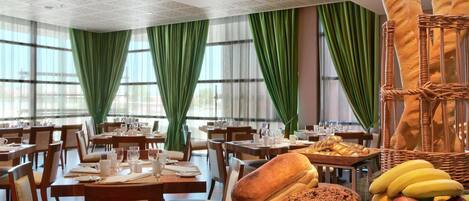A dining area with tables set for meals, green curtains, and a variety of breads and fruits on display.
