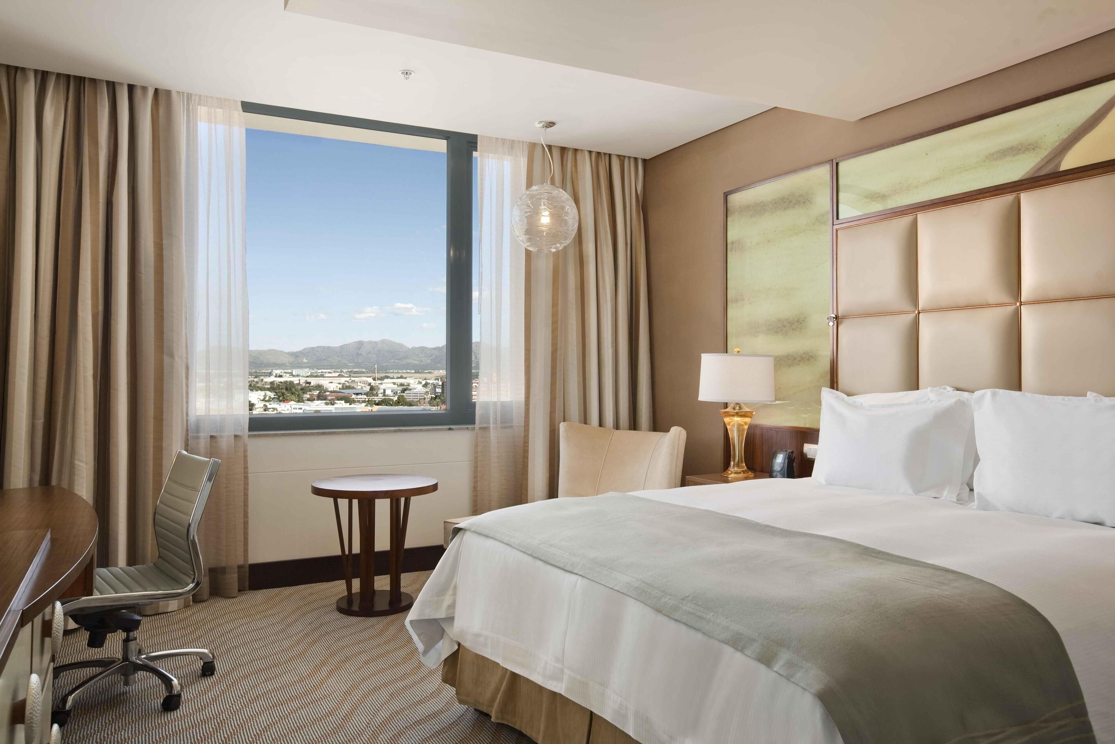 Habitación de hotel con una cama grande, un escritorio, una silla, una mesita y vistas a un paisaje urbano a través de un ventanal.