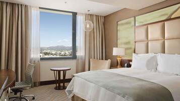 Habitación de hotel con una cama grande, un escritorio, una silla, una mesita y vistas a un paisaje urbano a través de un ventanal.