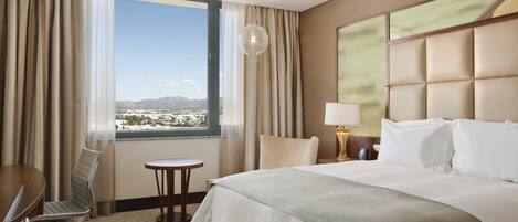 Une chambre d’hôtel avec un grand lit, un bureau, une chaise, une petite table et une vue sur le paysage urbain, visible à travers une grande fenêtre.