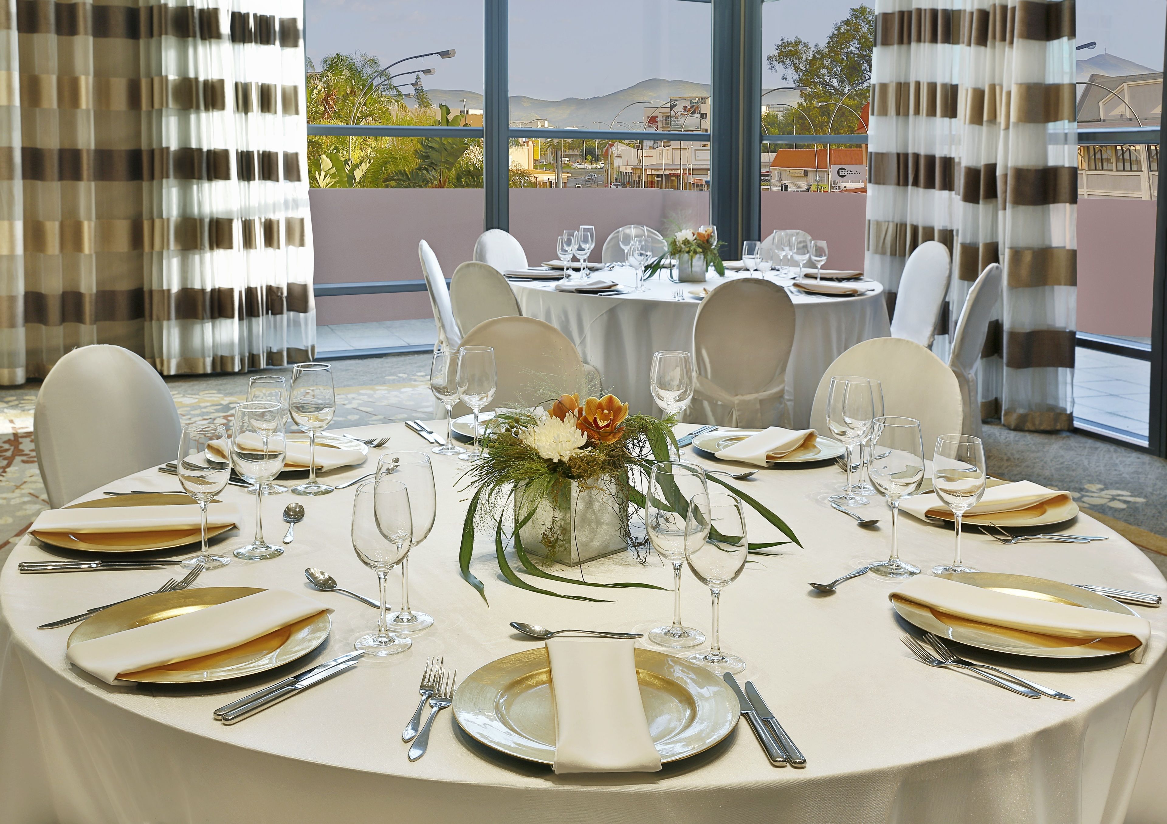 A round dining table set for a formal meal with white tablecloths, gold-rimmed plates, and clear glassware.