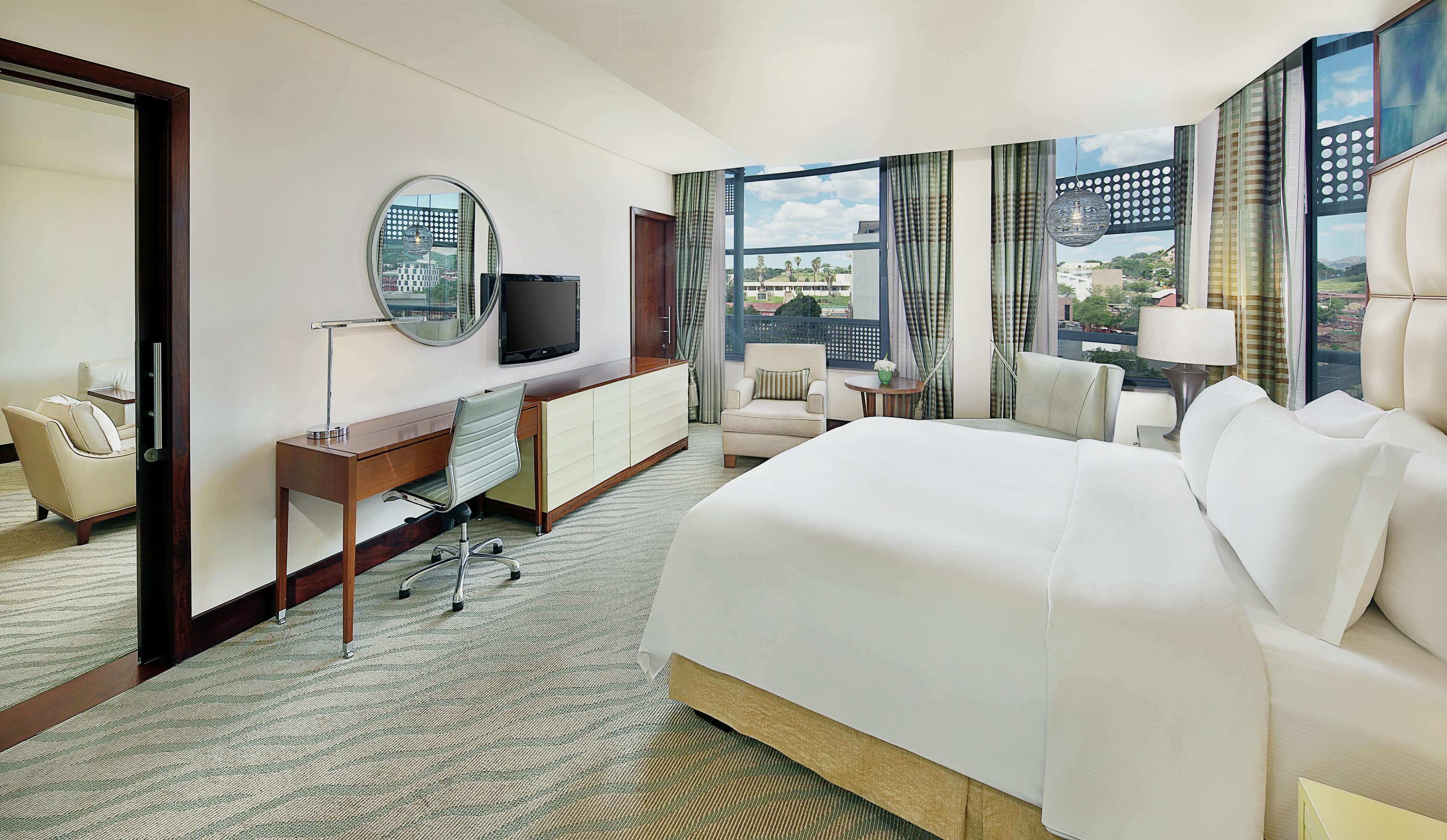 Habitación de hotel con una cama grande, un escritorio con silla, televisión y vistas a la ciudad a través de amplias ventanas.