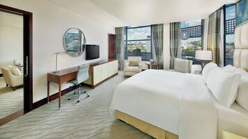 Habitación de hotel con una cama grande, un escritorio con silla, televisión y vistas a la ciudad a través de amplias ventanas.