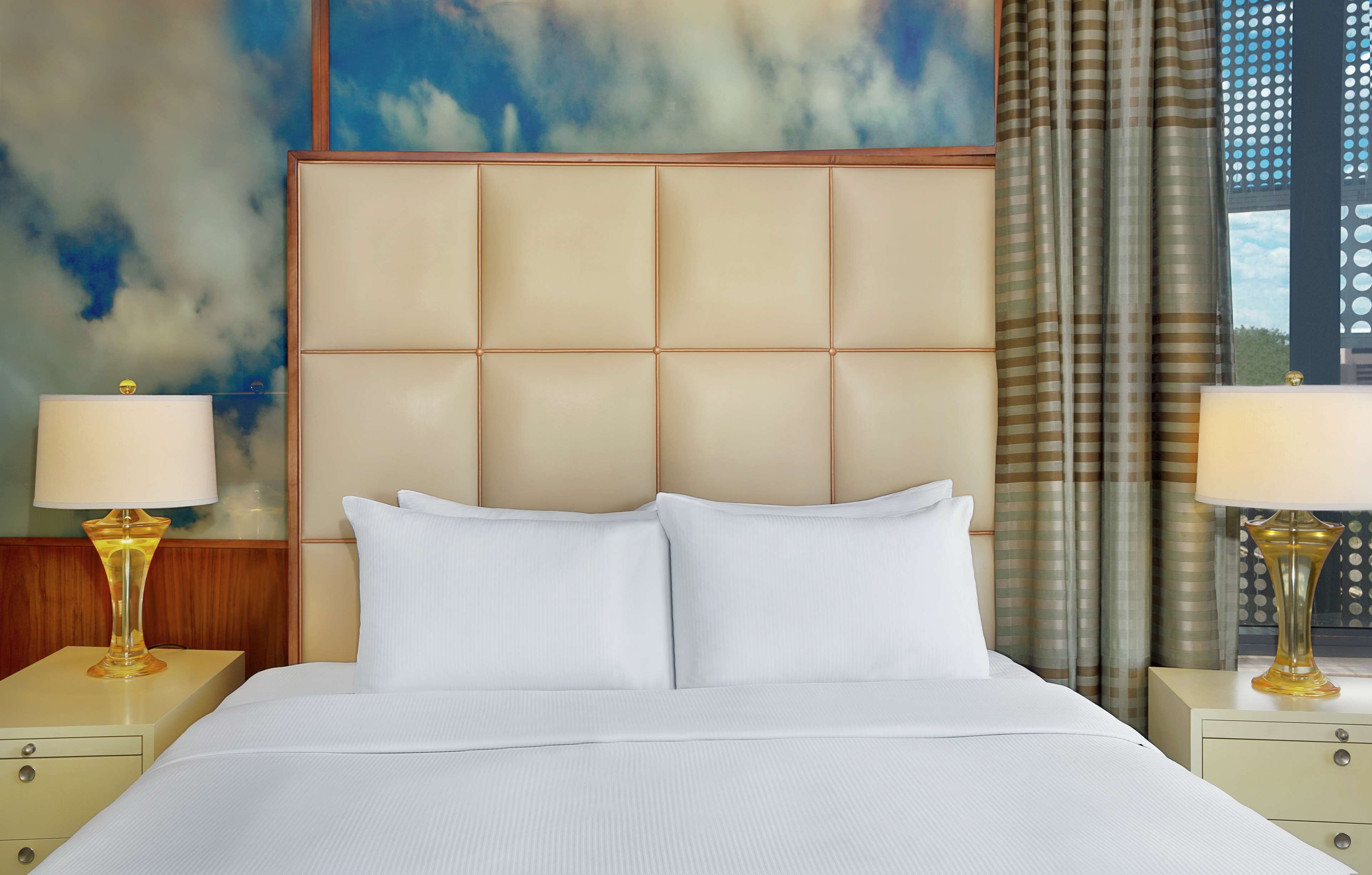 A hotel room with a large bed, two bedside tables with lamps, a headboard, and a window with curtains.