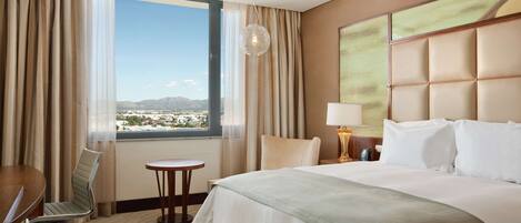 Habitación de hotel con una cama grande, un escritorio, una silla, una mesita y vistas a la ciudad desde la ventana.