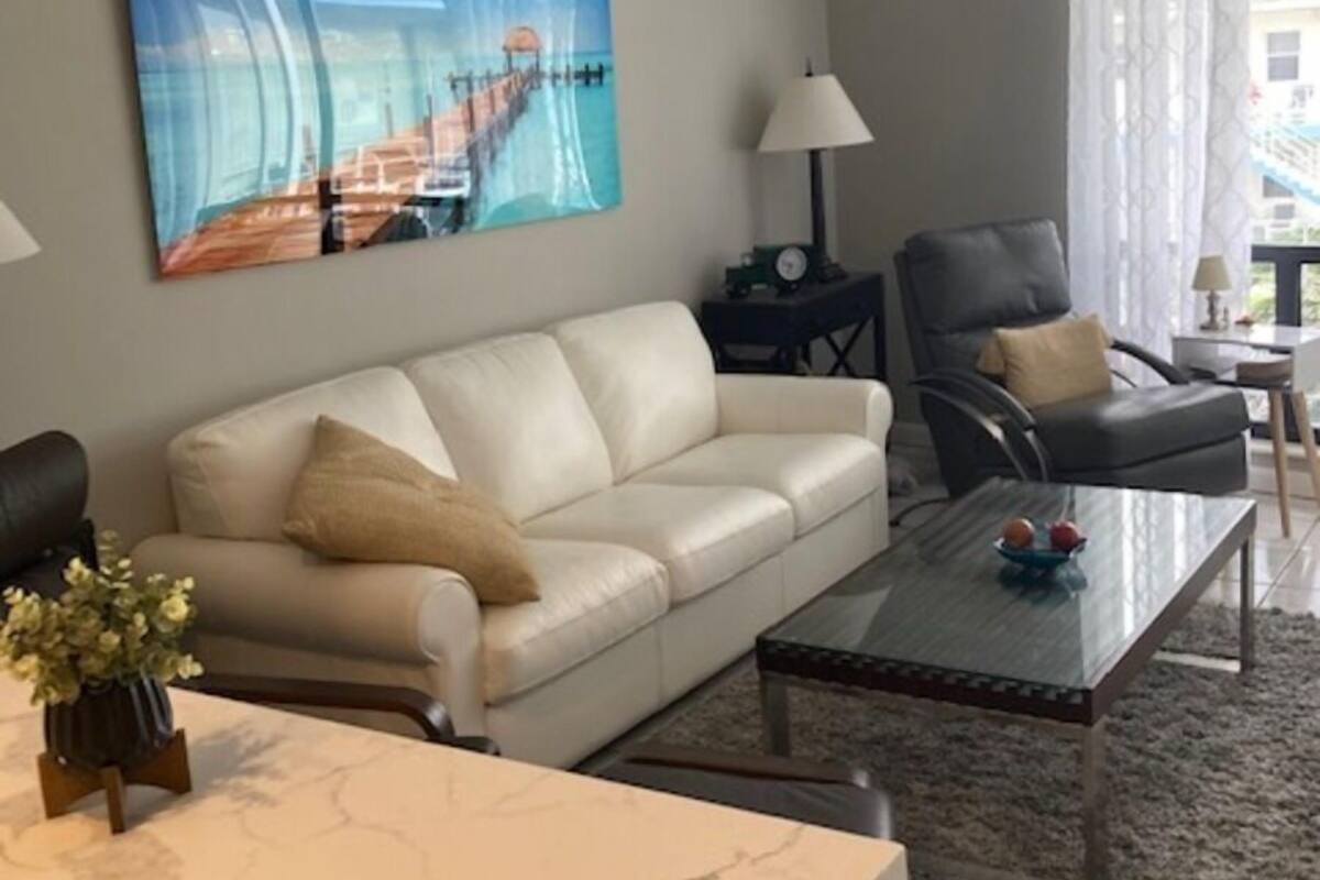 A living room with a white sofa, a glass coffee table, and a painting of a pier.