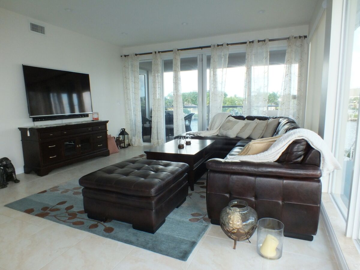 A modern living room with a large flat-screen TV, dark brown leather sofas, a glass coffee table, and a large window with curtains.