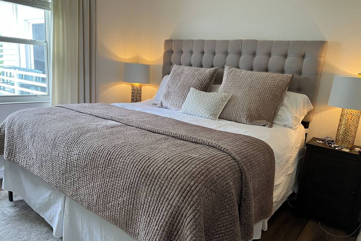 A neatly made bed with a grey headboard, a large beige blanket, and two bedside lamps.