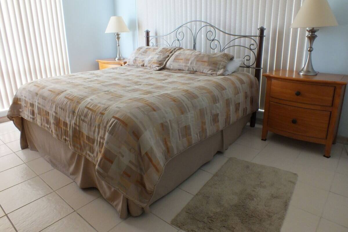 A bedroom with a bed, two bedside tables, a nightstand, a rug, and a window with blinds.