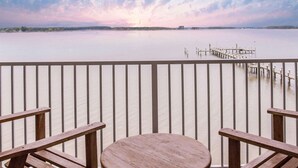 A wooden deck with a round table and benches, overlooking a calm body of water with a dock extending into it.