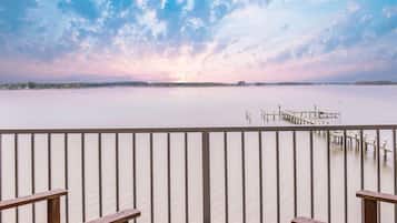 A wooden deck with a round table and benches, overlooking a calm body of water with a dock extending into it.