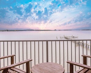 A wooden deck with a round table and benches, overlooking a calm body of water with a dock extending into it.