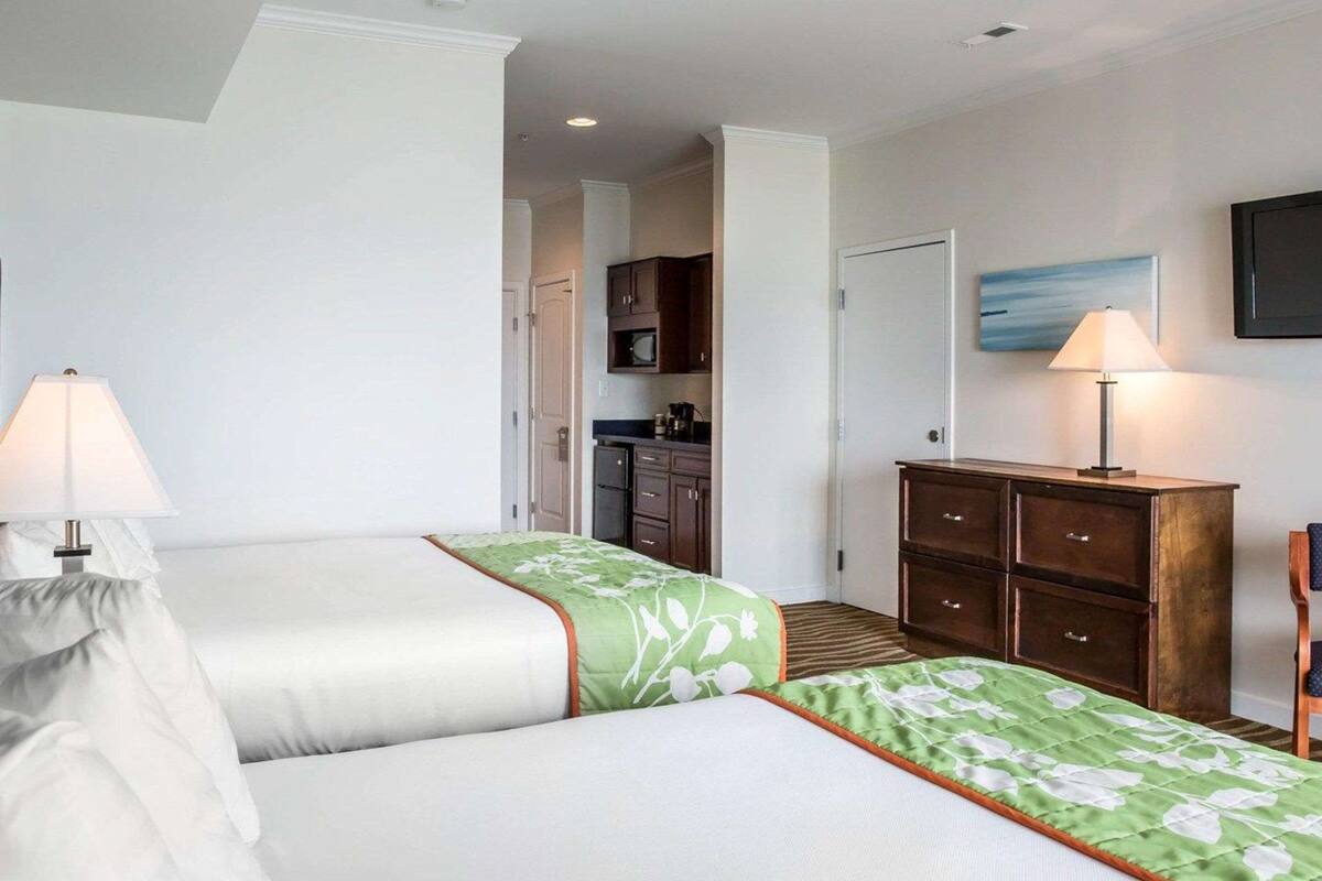 A hotel room with two beds, a kitchenette, a wooden dresser, and a painting on the wall.