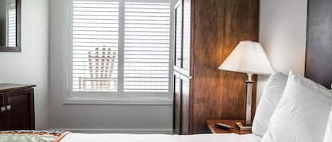 A hotel room with a large bed, a nightstand with a lamp, a wooden wardrobe, and a window with blinds.
