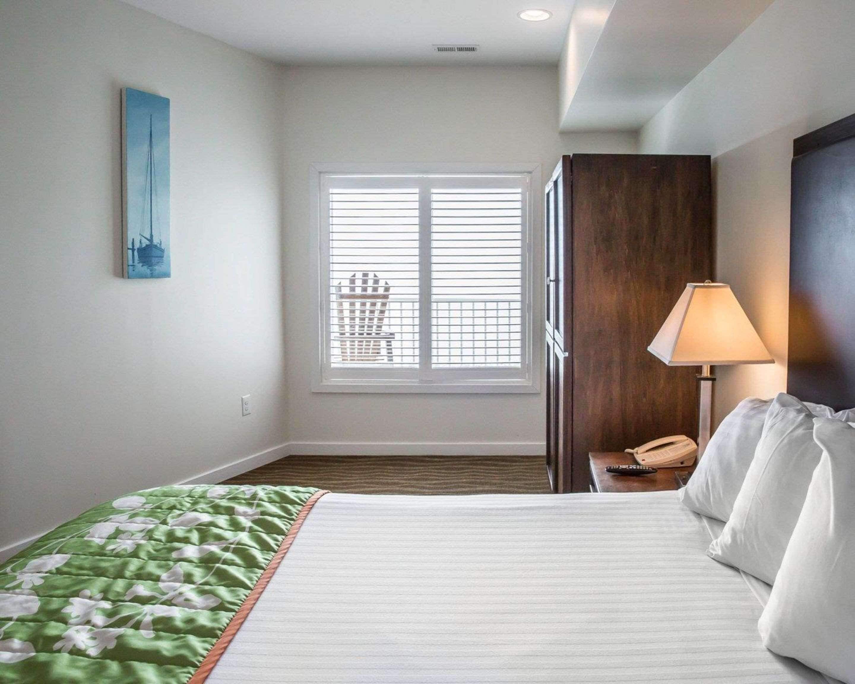 A bedroom with a bed, a nightstand, a lamp, a window with blinds, and a painting on the wall.
