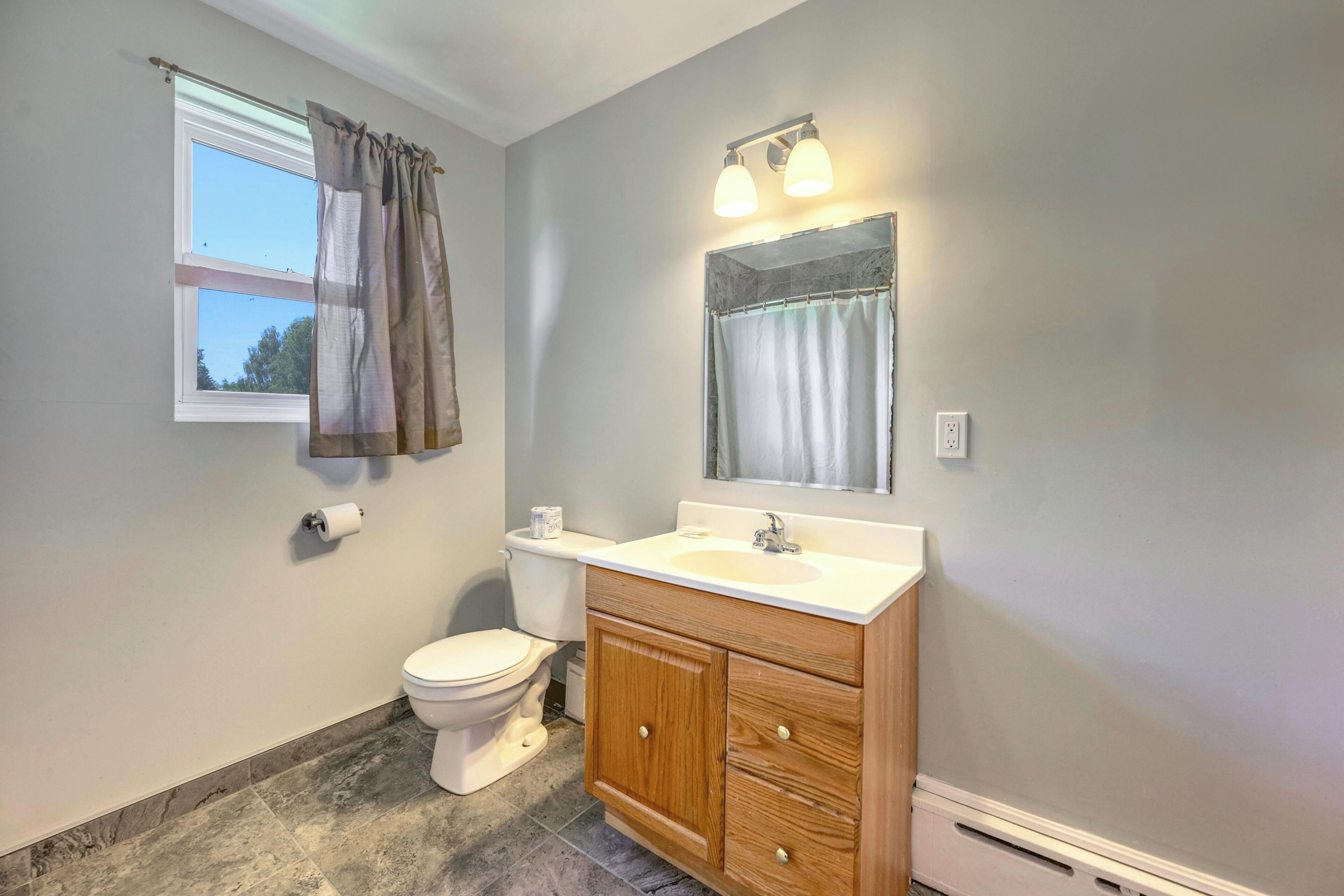 A bathroom with a toilet, sink, vanity, and a window with curtains.