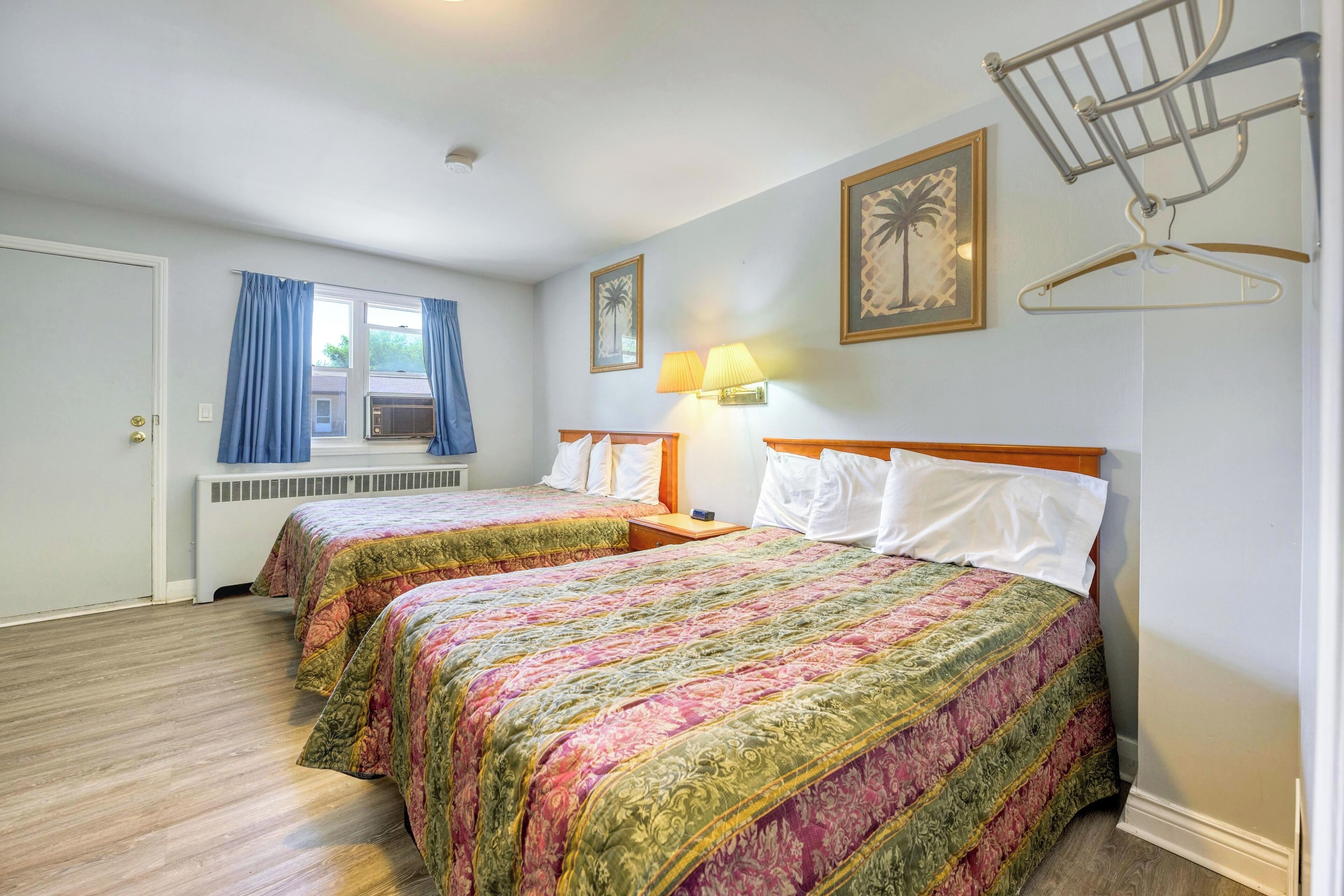 A hotel room with two beds, a window with blue curtains, a wall-mounted lamp, and a framed picture.