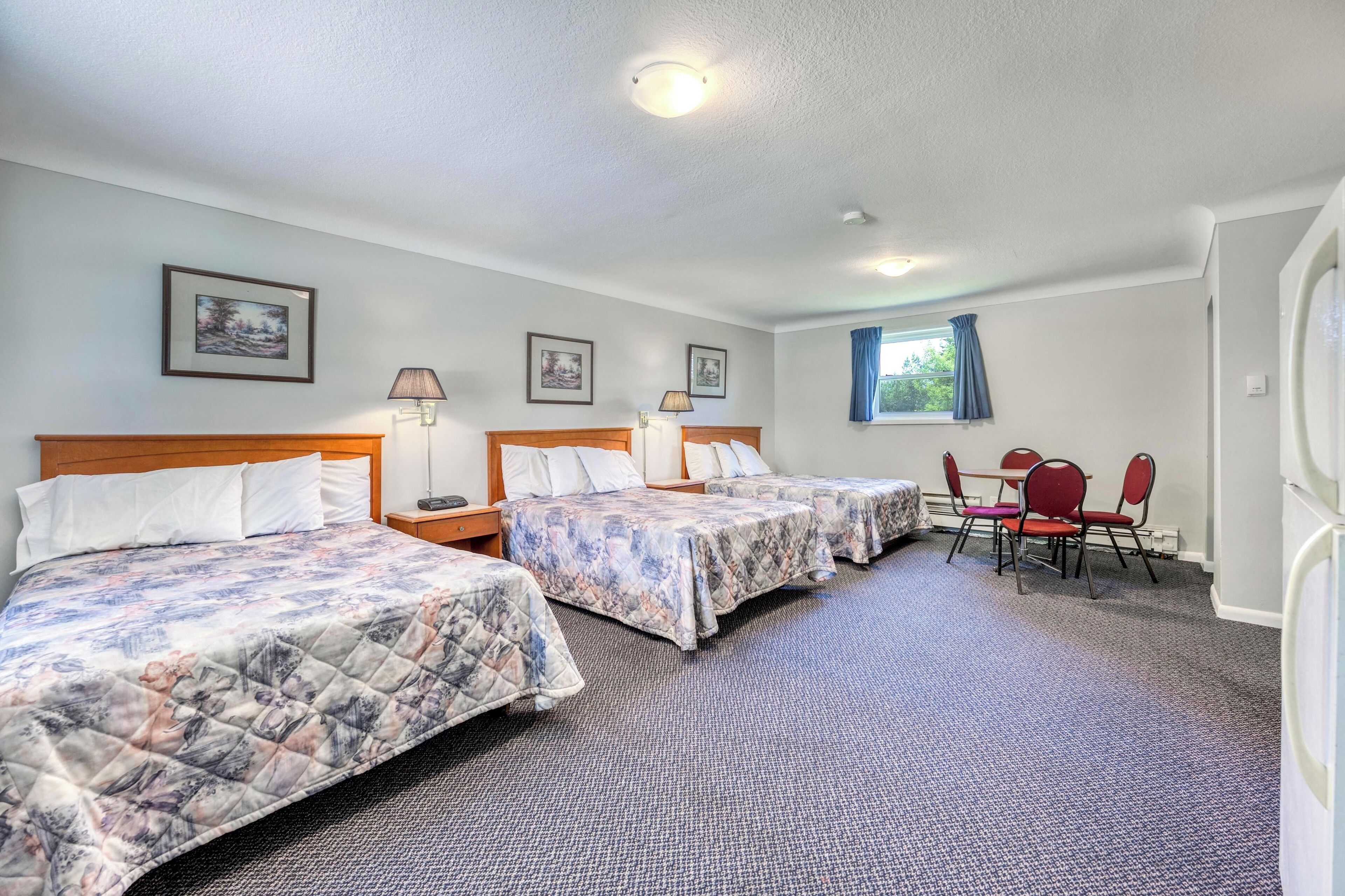 Una habitación de hotel con tres camas, una mesa de comedor y sillas, y una ventana con cortinas.