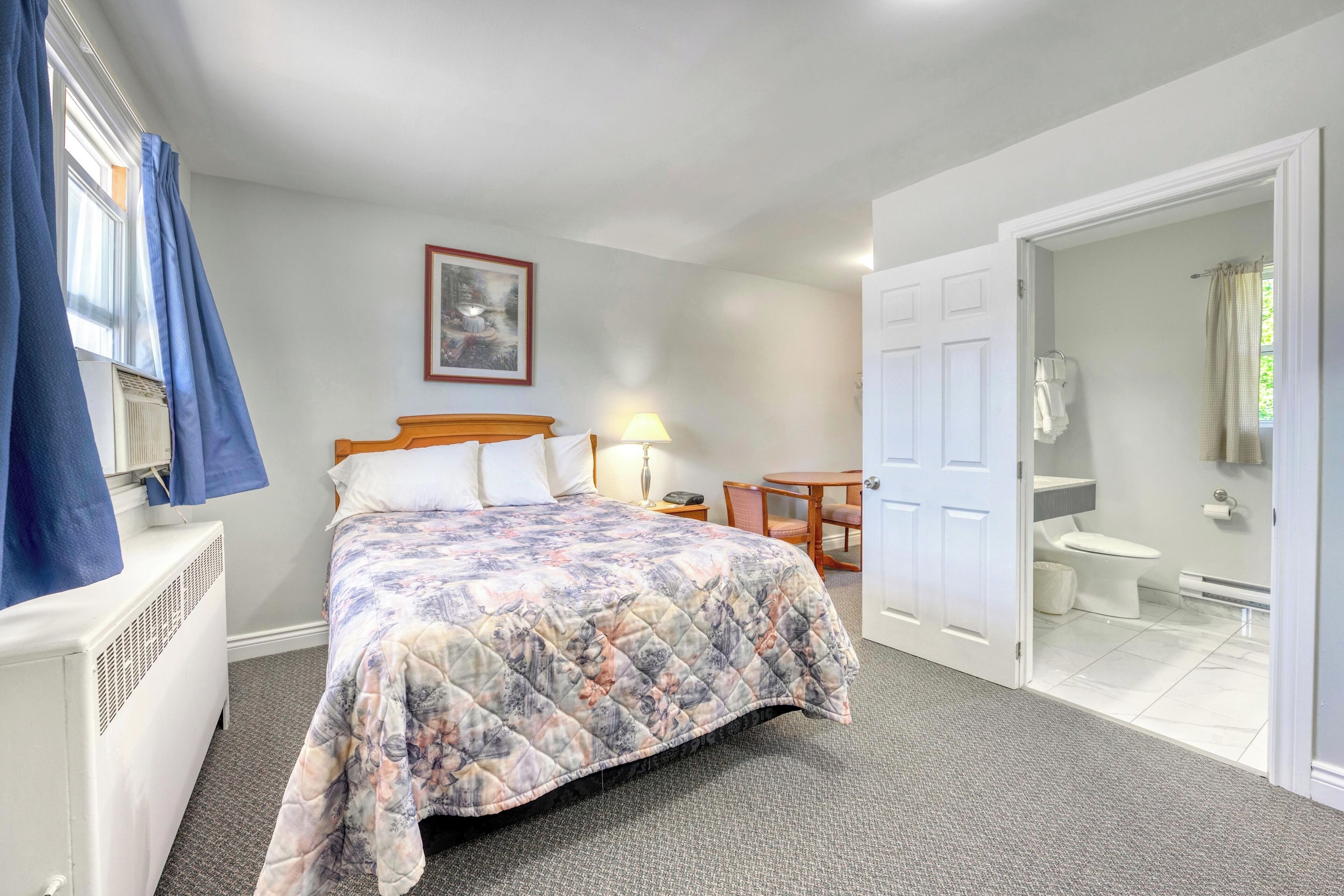 A bedroom with a bed, a bedside table, a lamp, a chair, and a bathroom visible through an open door.