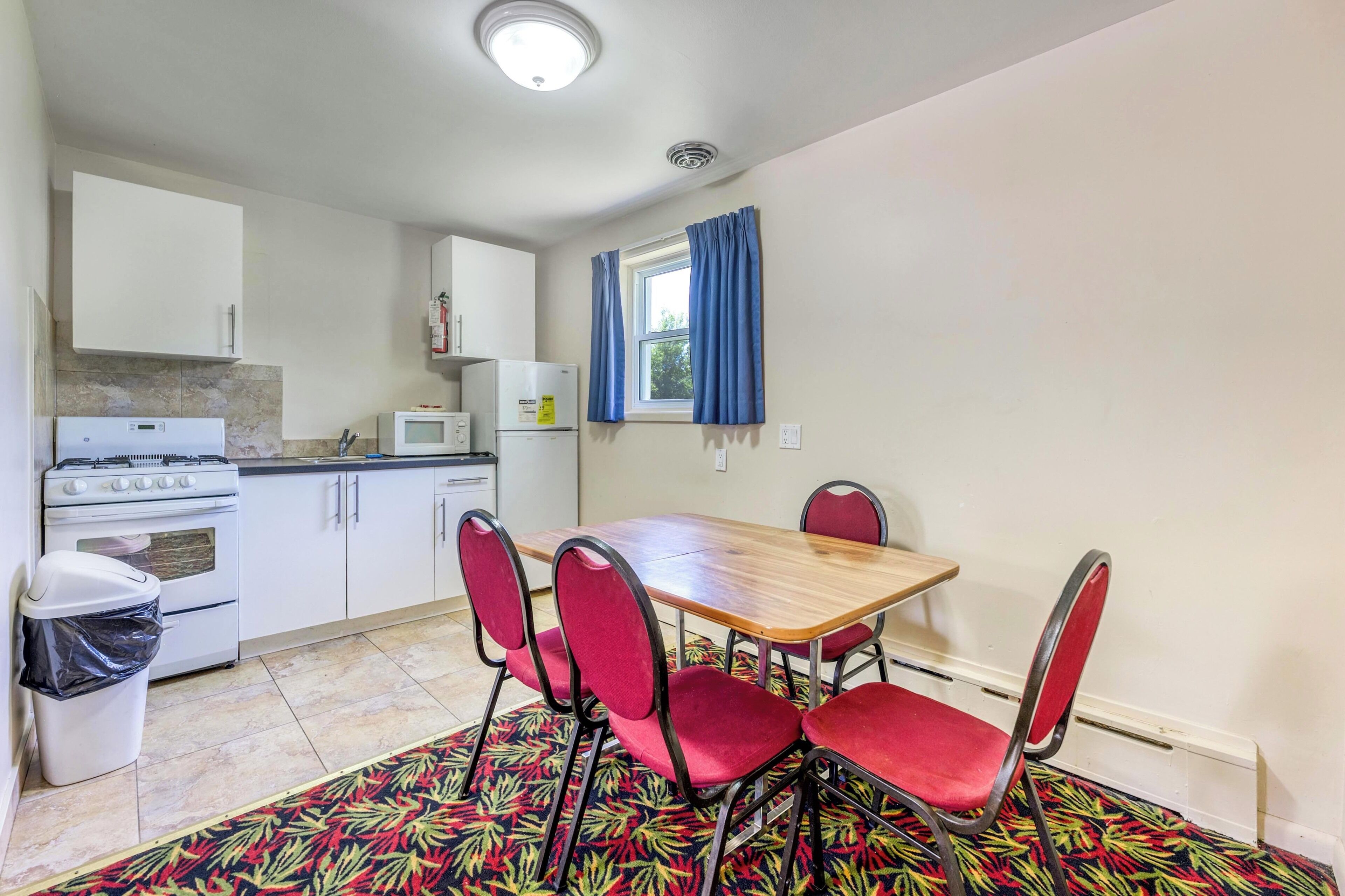 A compact kitchen with white cabinets, a stove, a microwave, and a window with blue curtains.