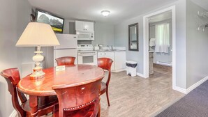 A dining area with a round table and chairs, a kitchen with white cabinets, a microwave, and a television on the wall. - The Gables inn (Sarnia)