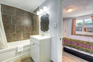 A bathroom with a bathtub, sink, and mirror. There is a window with curtains and a bed with a patterned bedspread.