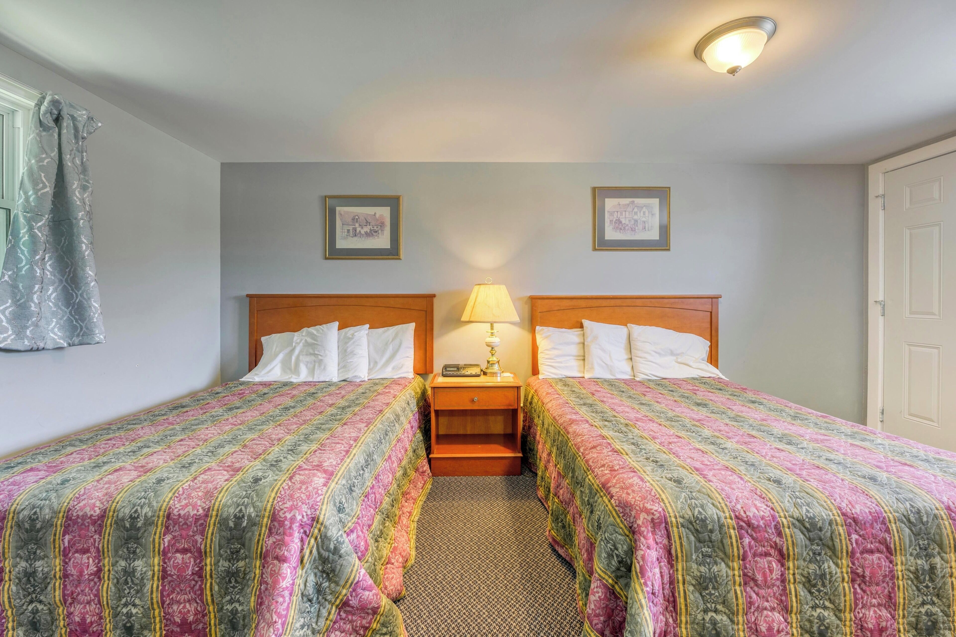 A hotel room with two single beds, a nightstand with a lamp, and a door.