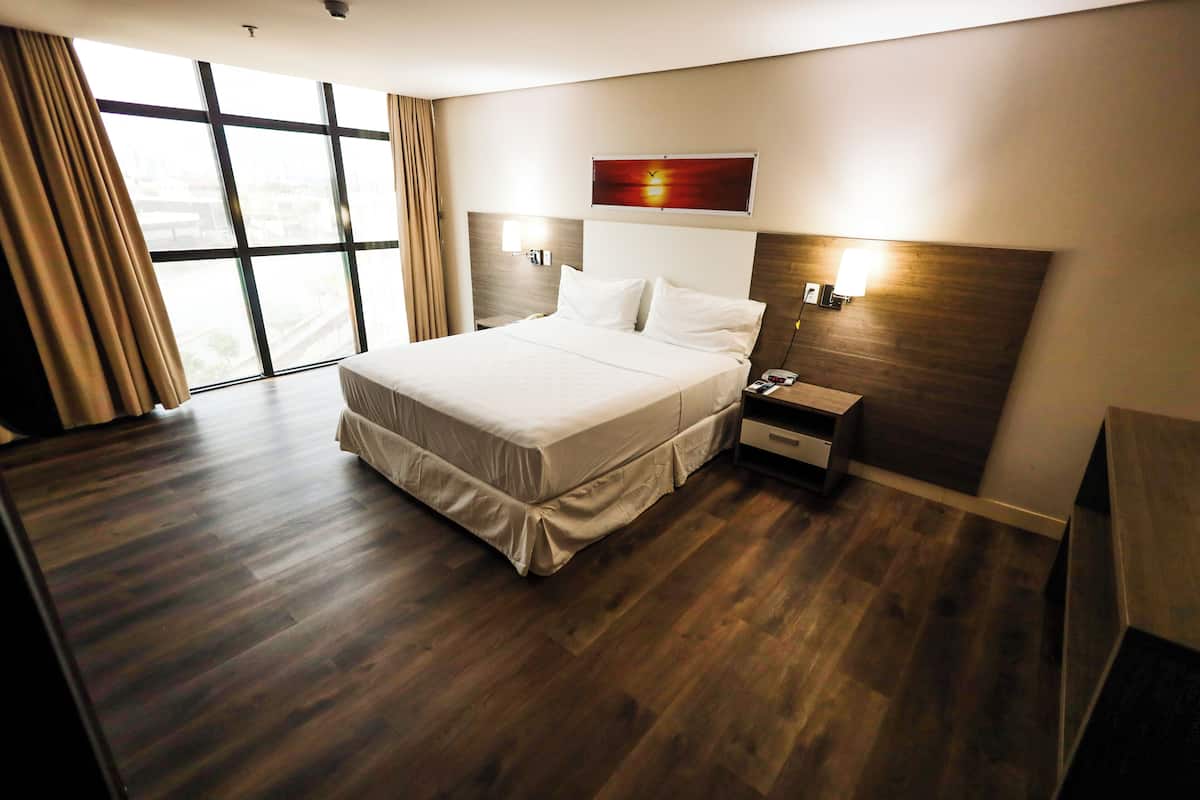 A modern hotel room with a large bed, wooden flooring, a nightstand, a wall-mounted lamp, and a large window with curtains.