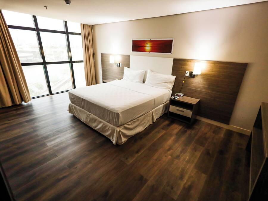 A modern hotel room with a large bed, wooden flooring, a nightstand, a wall-mounted lamp, and a large window with curtains.