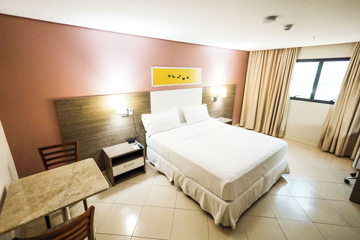 A hotel room with a bed, a desk, a chair, a nightstand, and a window with curtains.