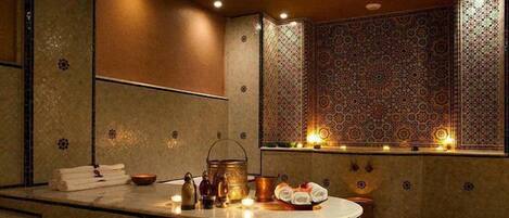 Une salle de bain avec un meuble-lavabo en marbre aux lignes courbes, un évier et un miroir. On y trouve des bougies sur le meuble-lavabo et un mur recouvert de carreaux à motifs.