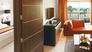 Een hotelkamer met een bed, een slaapbank, een salontafel met fruit, een televisie en een balkon met uitzicht op het strand.