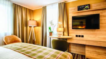 A hotel room with a bed, a desk, a TV, a lamp, curtains, and a plant.