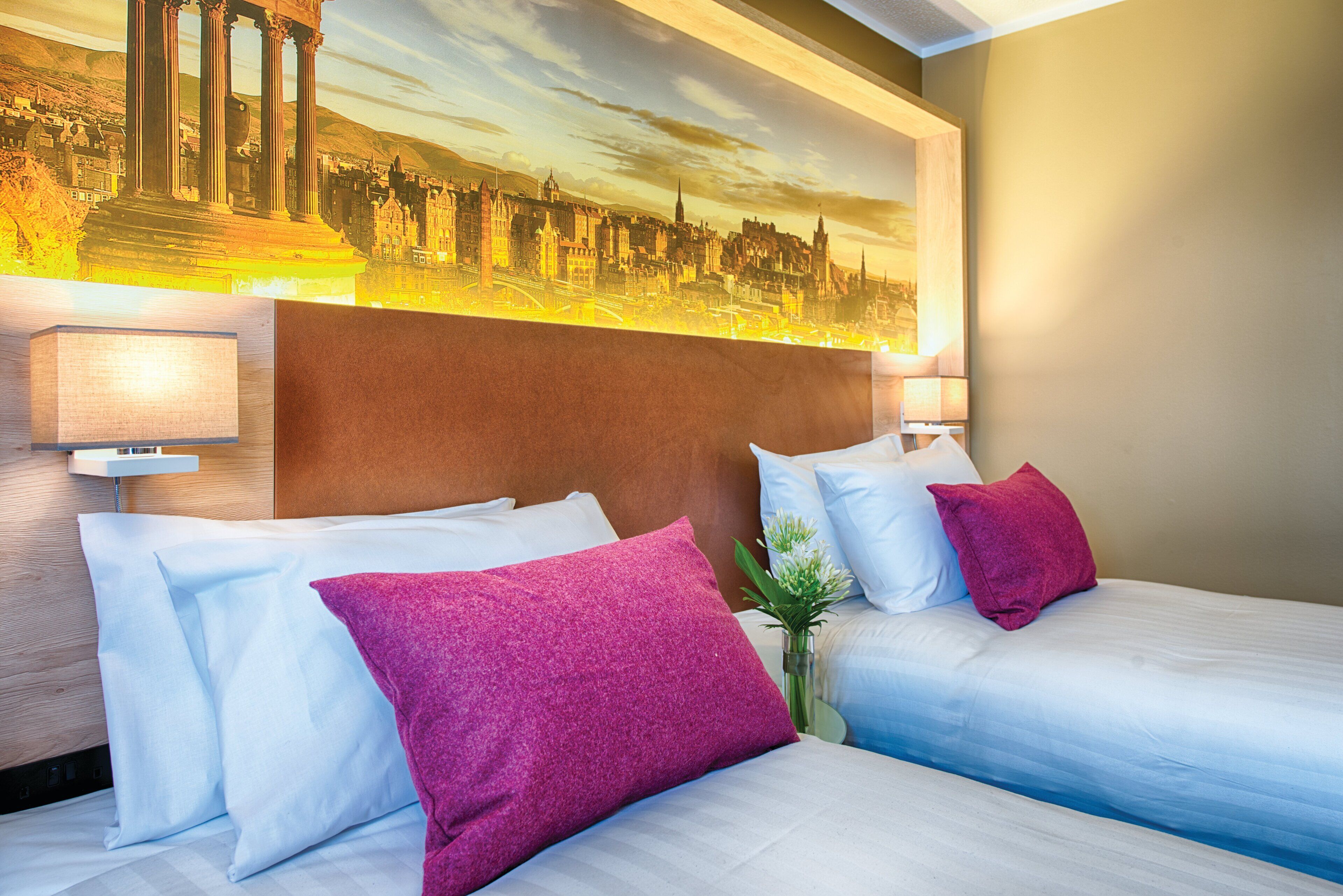 A hotel room with two beds, a large painting of a cityscape, and a modern headboard.