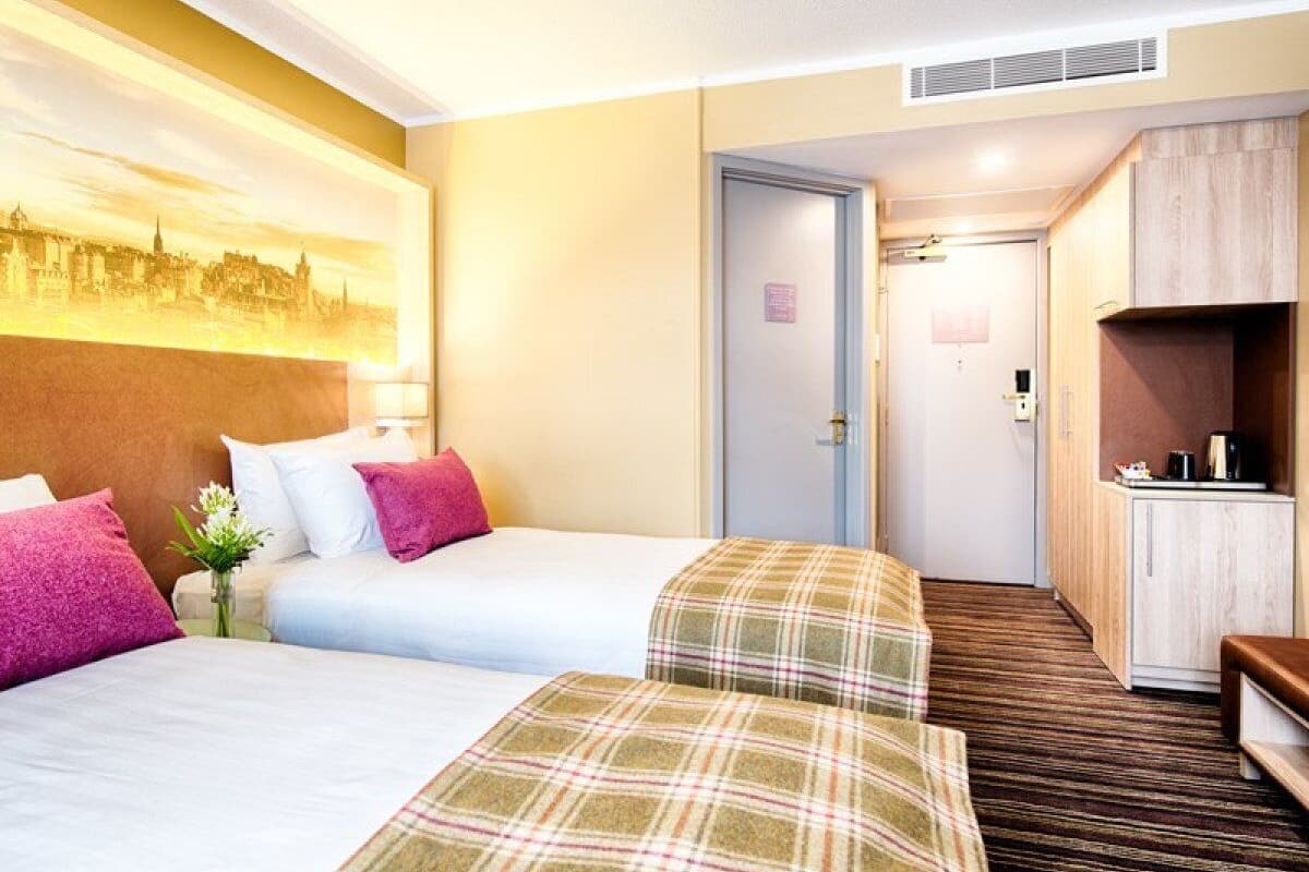 A hotel room with two beds, a small desk, a kitchenette, and a door leading to another room.