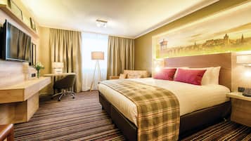 A hotel room with a bed, a desk, a chair, a television, and a cityscape painting on the wall.