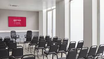 Un salón de conferencias con filas de sillas negras, un letrero rojo en la pared y ventanas amplias.