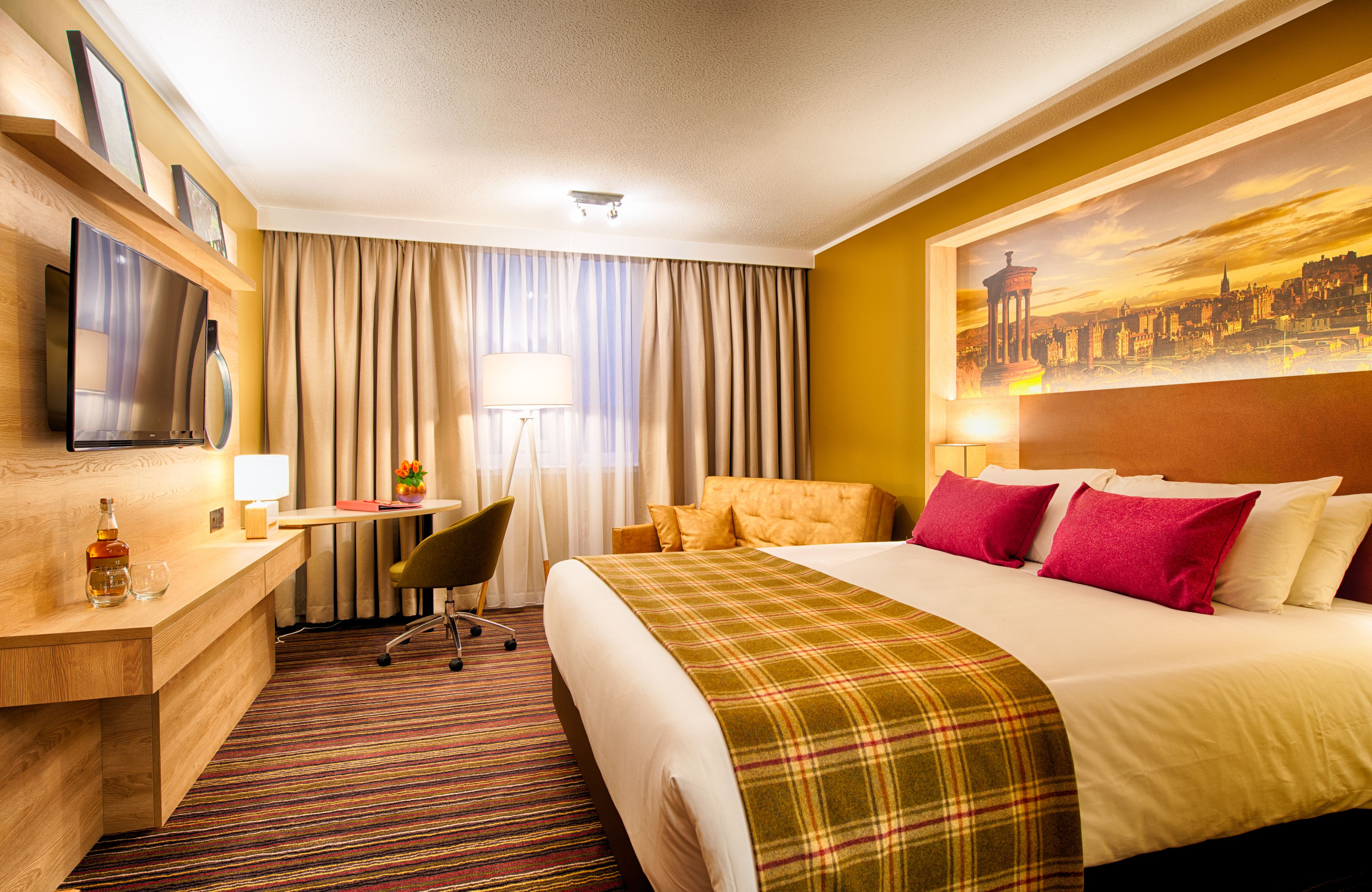 A hotel room with a bed, a desk, a chair, a television, and a painting on the wall.