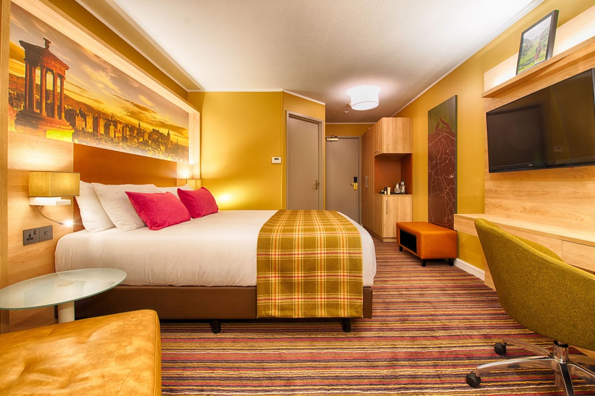A hotel room with a bed, a small table, a chair, a television, and a painting on the wall.
