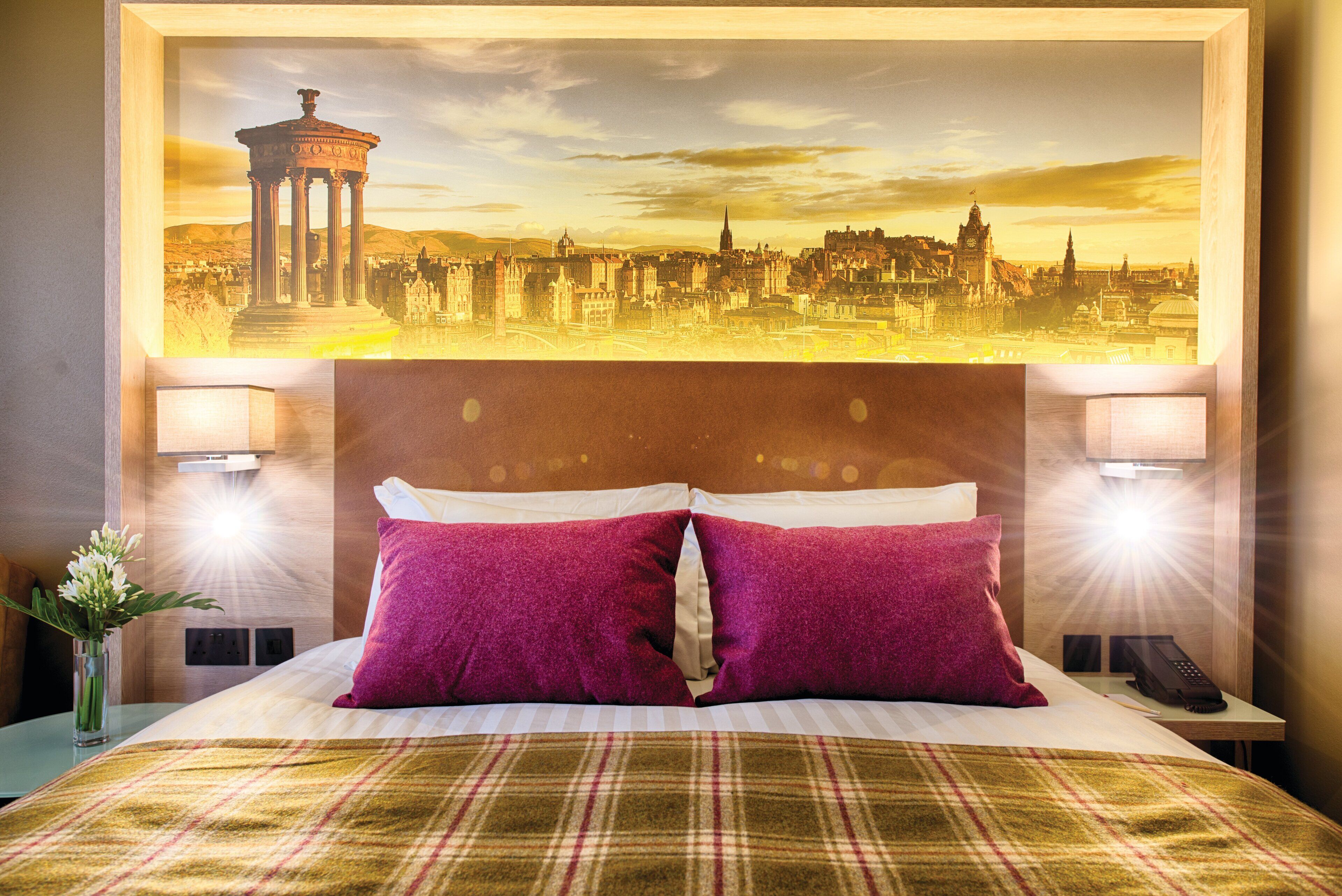 Ein Hotelzimmer mit einem Bett, zwei Kissen, einem Nachttisch, einer Lampe und einem an der Wand hängenden Stadtbild.