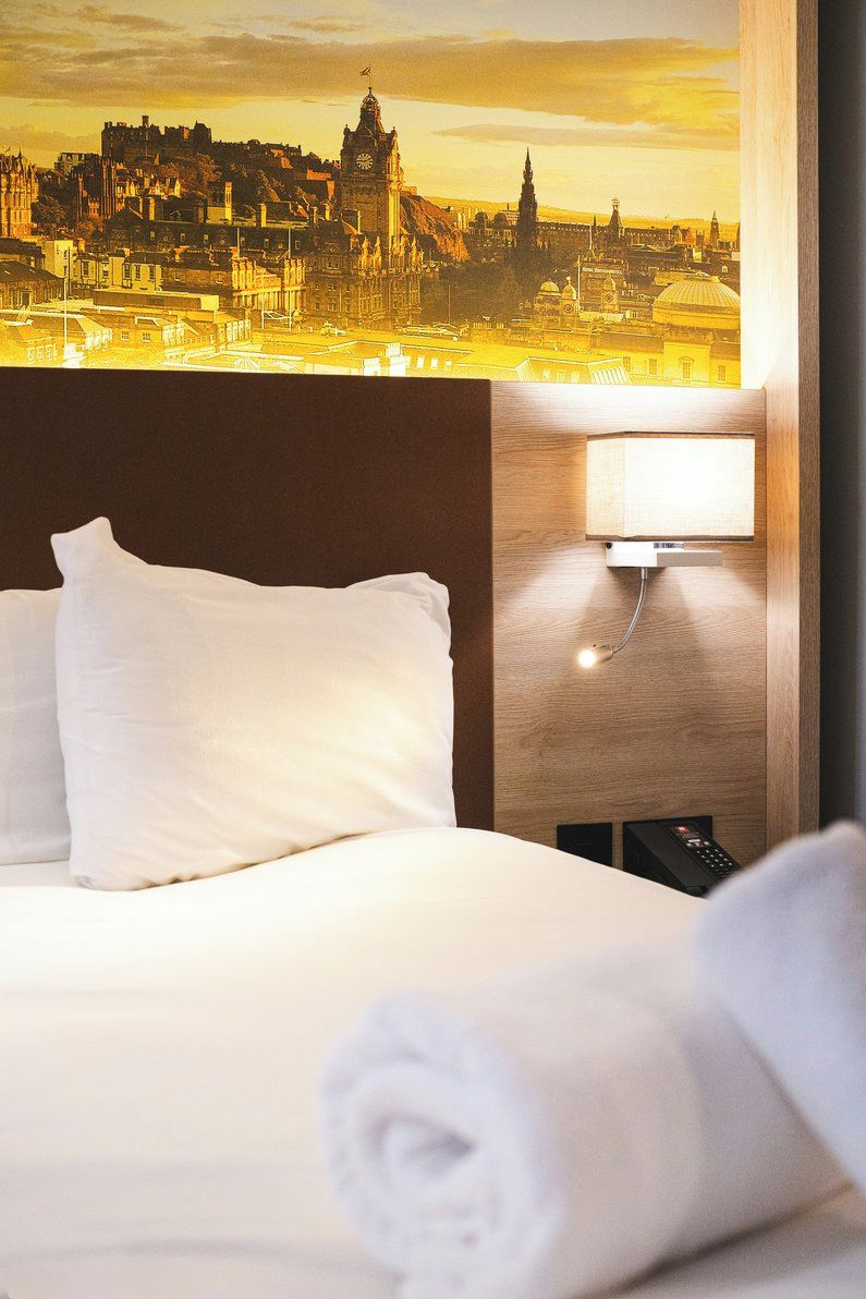 A hotel room with a bed, a nightstand, a lamp, and a view of a cityscape.