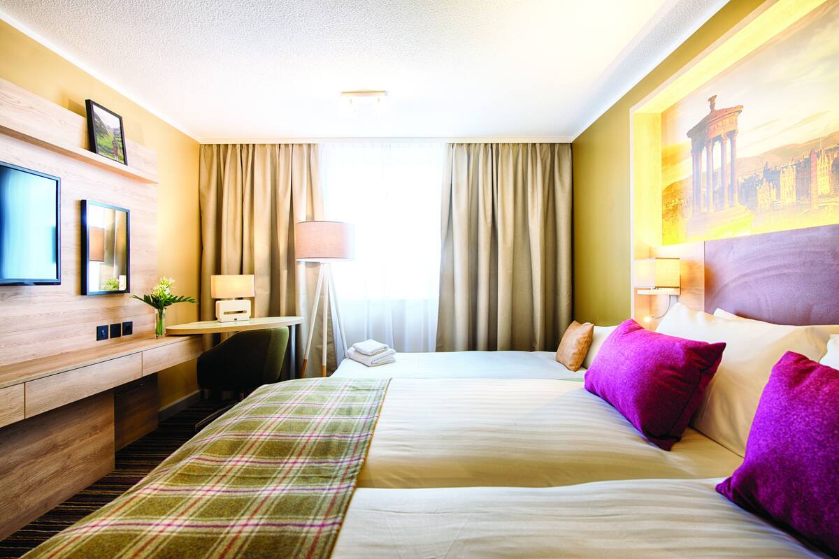 A hotel room with a bed, a desk, a chair, a TV, and a painting on the wall.