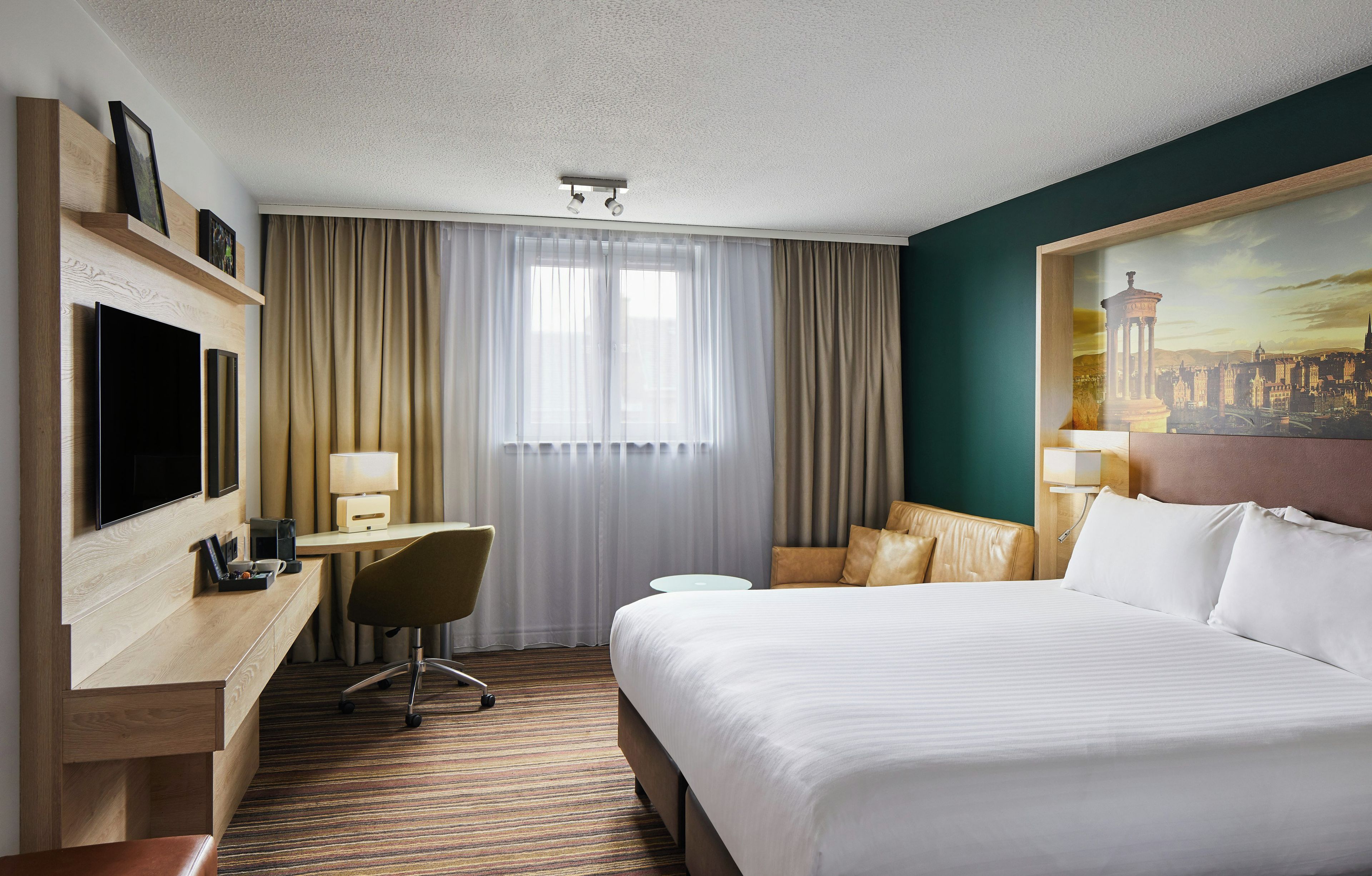 A hotel room with a bed, desk, chair, television, and a painting on the wall.