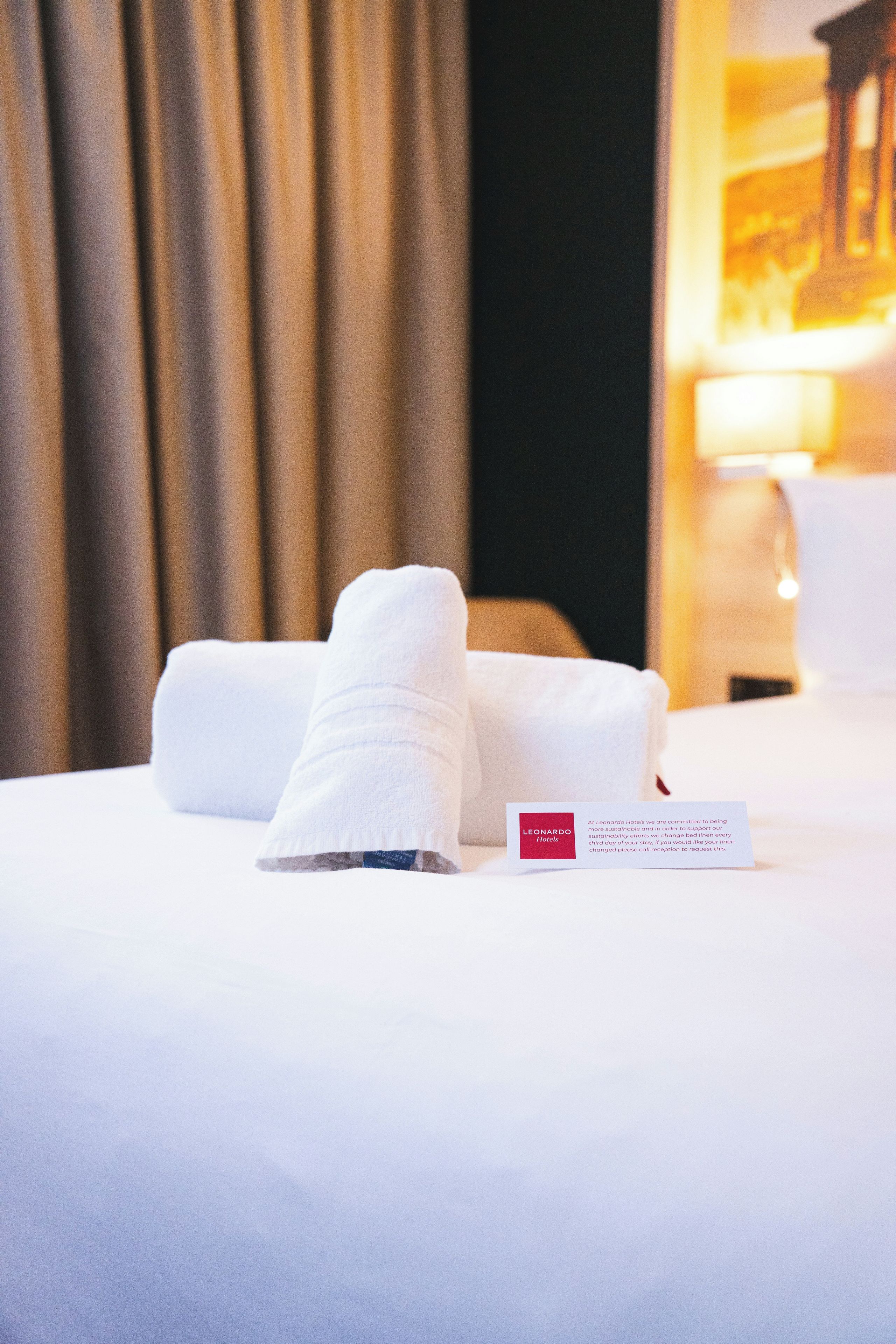 Un lit bien fait, avec du linge de lit blanc et une serviette pliée. Une enseigne portant le logo de l’hôtel est visible.