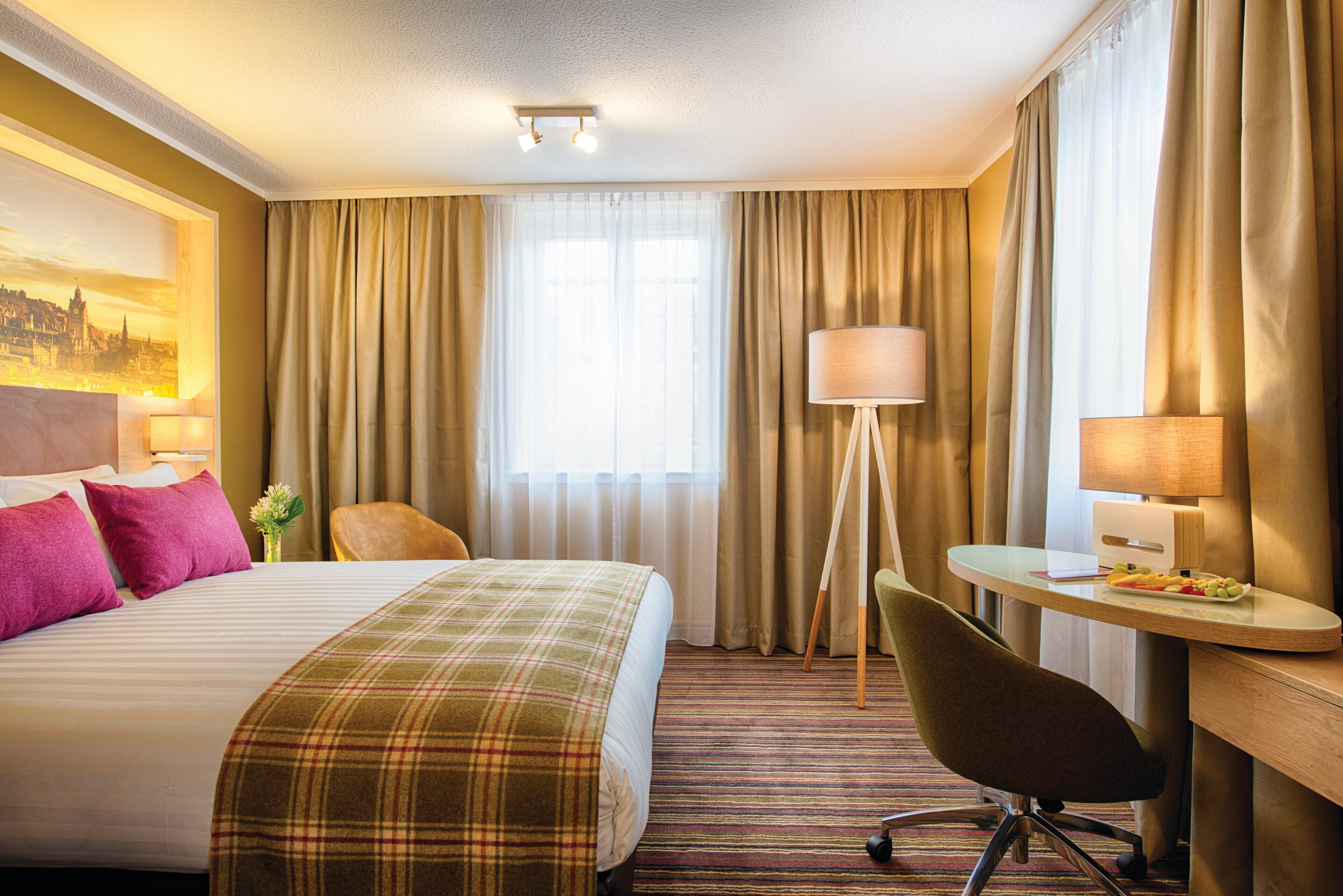 Ein Hotelzimmer mit Bett, Schreibtisch, Stuhl, Lampe und einem fensterbepflanzten Fenster.